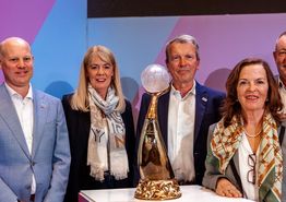 Vertreter und Vertreterinnen der Austragungsstätten stehen um den Pokal herum und lachen in die Kamera.
