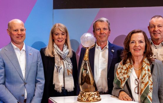 Vertreter und Vertreterinnen der Austragungsstätten stehen um den Pokal herum und lachen in die Kamera.