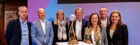 Vertreter und Vertreterinnen der Austragungsstätten stehen um den Pokal herum und lachen in die Kamera.