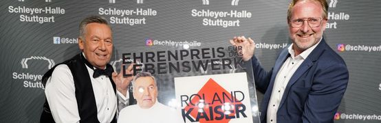 Roland Kaiser und René Otterbein halten den Ehrenpreis in Form einer Plexiglasplatte und lachen in die Kamera.