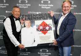 Roland Kaiser und René Otterbein halten den Ehrenpreis in Form einer Plexiglasplatte und lachen in die Kamera.