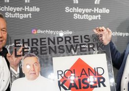 Roland Kaiser und René Otterbein halten den Ehrenpreis in Form einer Plexiglasplatte und lachen in die Kamera.